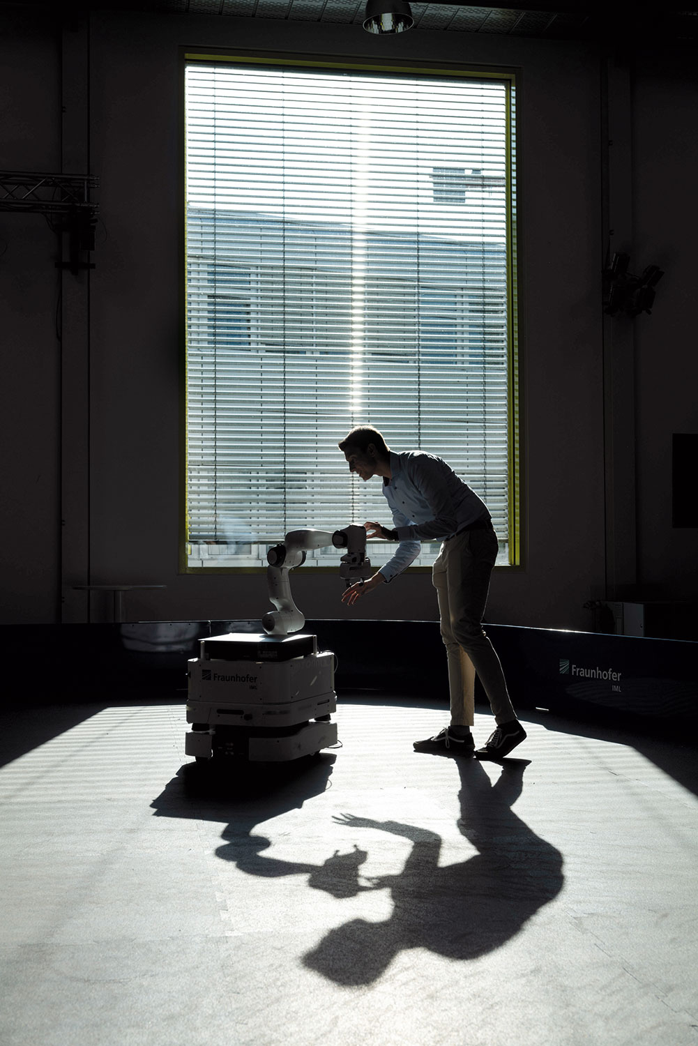 Informatiker Sebastian Hoose in einem  Institutsraum mit halb abgedunkelter Jalousie an einem großen Fenster und hantiert an dem transportablen Roboter-System RAI - Remote AI.