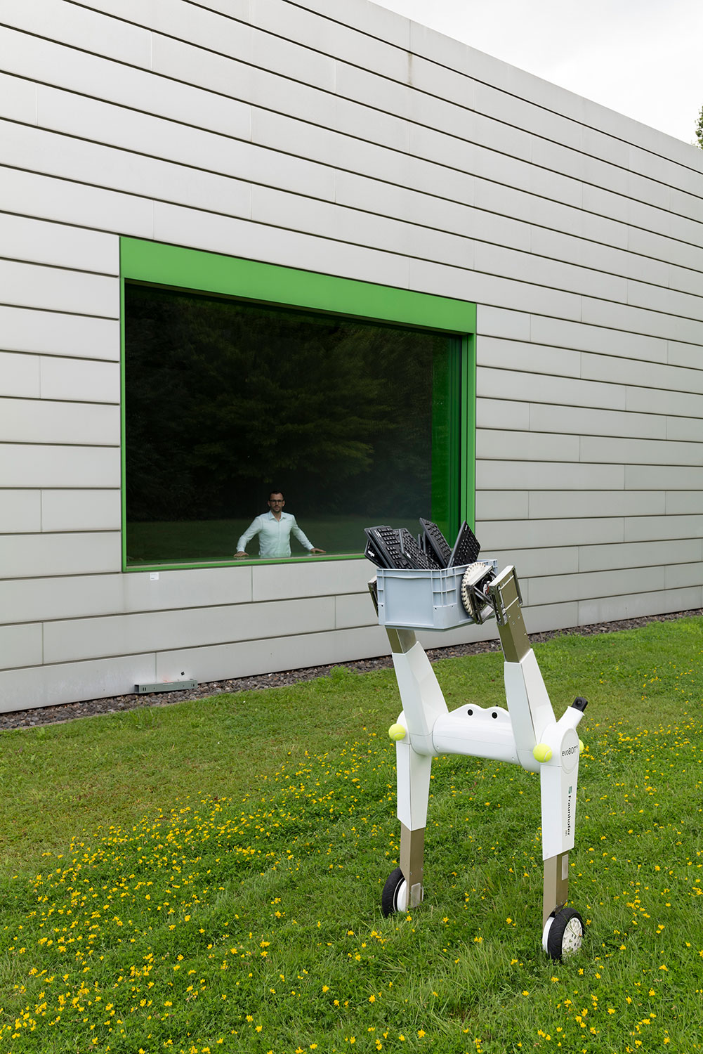 Der Forscher Leon Siebel-Achenbach steht im Inneren des Institutsgebäudes am Fenster und blickt auf den Serviceroboter Evobot, der auf einer Wiese steht und einem Transportkorb mit Computer-Tastaturen hoch hält.