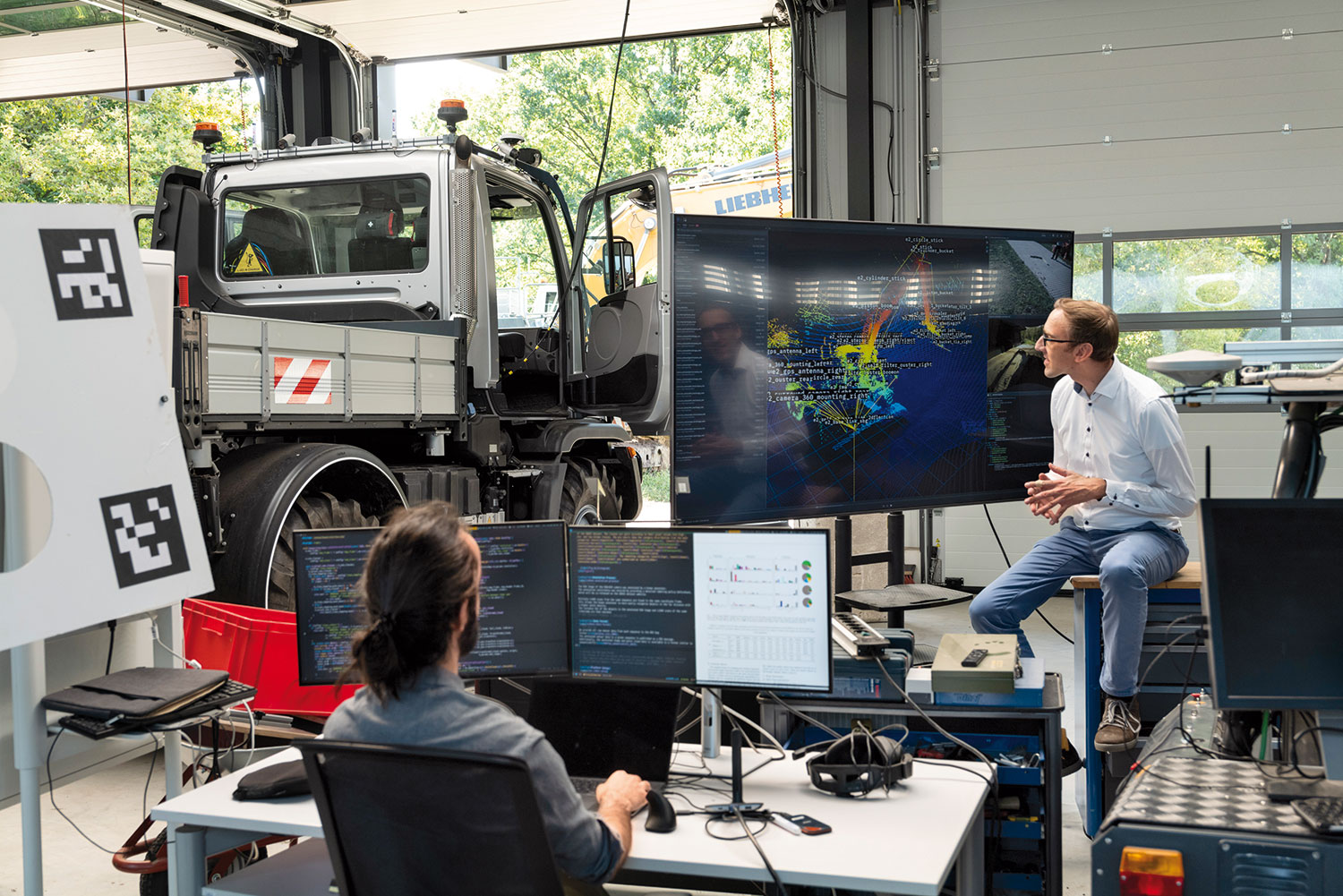 Forscher Janko Petereit sitzt im Institutsgebäude vor einem Bildschirm mit einer bunten Grafik eines Raupenbaggers, dahinter ein Unimog in der Ausfahrt, vor ihm ein Mitarbeiter vor zwei kleineren Bildschirmen.