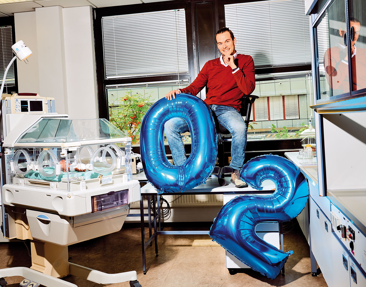 Dr. Felix Wiegandt sitzt in einem Labor erhöht auf einem Tisch, vor ihm 2 aufblasbare Luftballons O2 für Sauerstoff, daneben steht ein Frühchen-Brutkasten, darin ein täuschend echtes Neugeborenes aus Kunststoff.