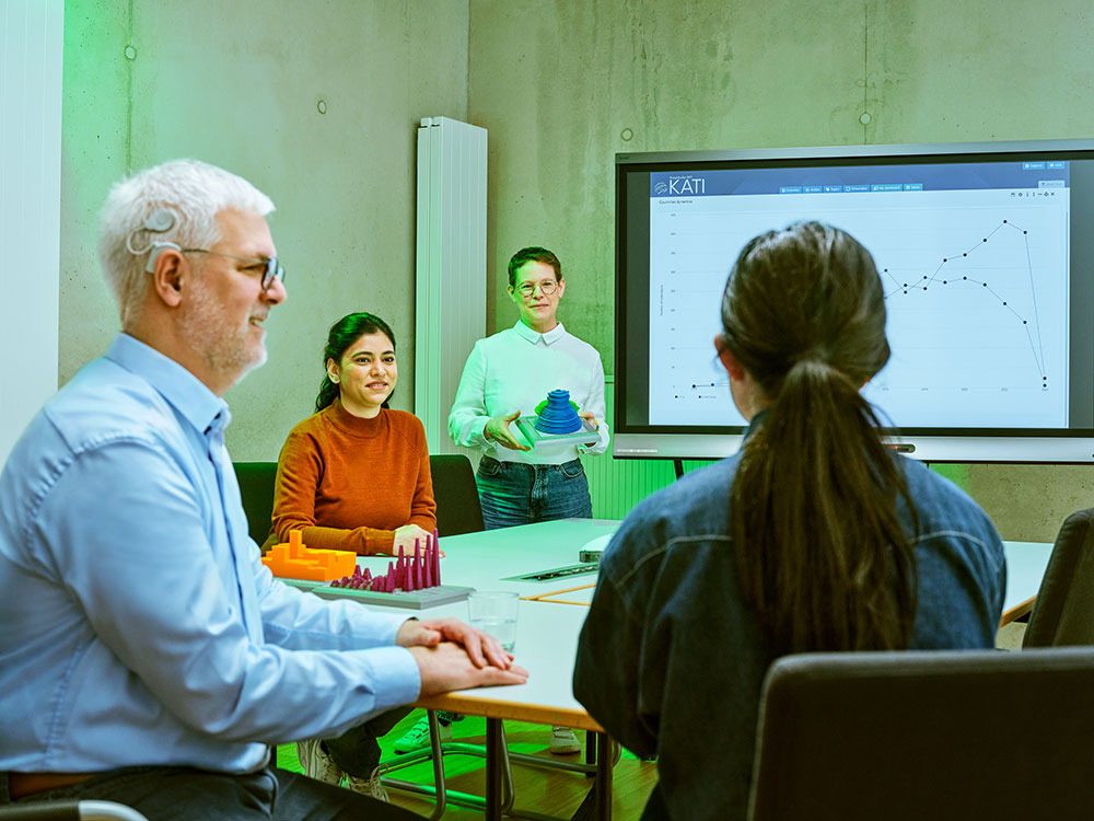Symbolbild Inklusion: Mitarbeitende in einem Besprechungsraum: Eine Person mit Hörimplantat sitzt im Vordergrund. Im Hintergrund präsentieren Teammitglieder Modelle und Diagramme auf einem Bildschirm.