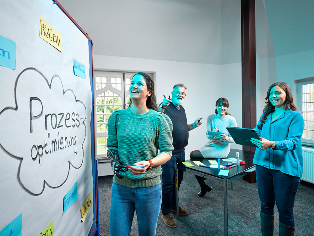 Symbolbild Inklusion: Gruppe von fünf Mitarbeitenden bei einem Workshop. Eine Frau mit Armprothese steht vor einem Flipchart mit der Aufschrift 'Prozessoptimierung', während die anderen Personen diskutieren und Materialien wie Tablets und Moderationskarten nutzen.
