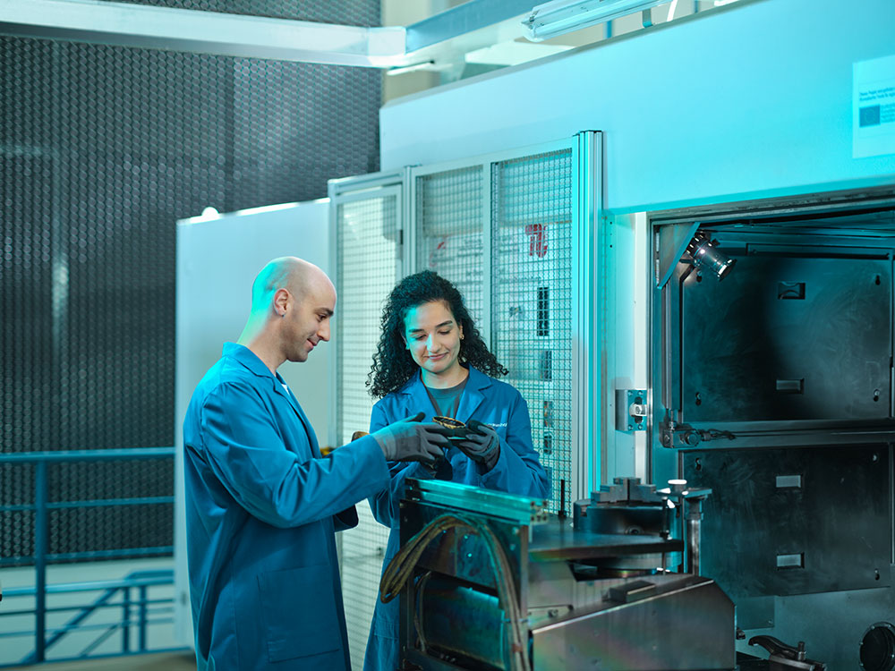 Symbolbild Gender: Eine weibliche und ein männlicher Mitarbeitende in blauen Laborkitteln arbeiten gemeinsam an einem technischen Gerät in einer Werkstattumgebung. Beide blicken auf ein Bauteil in ihren Händen.