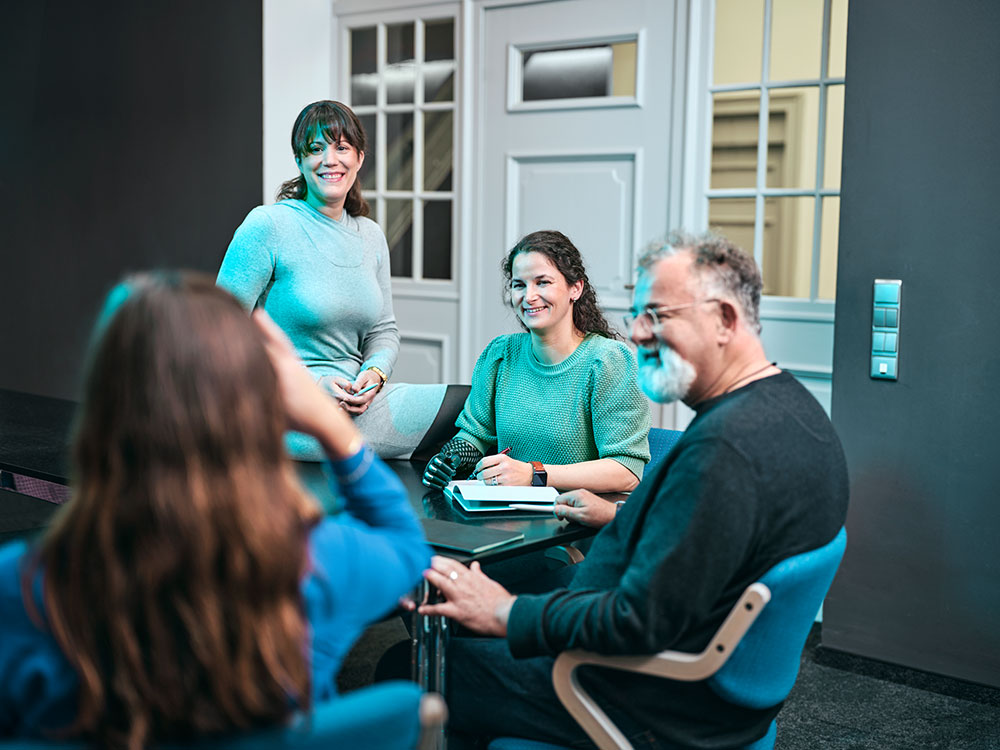 Symbolbild Inklusion: Gruppe von vier Mitarbeitenden in einem Besprechungsraum. Drei Personen sitzen um einen Tisch, während eine weitere Person am Tisch steht. Alle blicken einander freundlich zu.