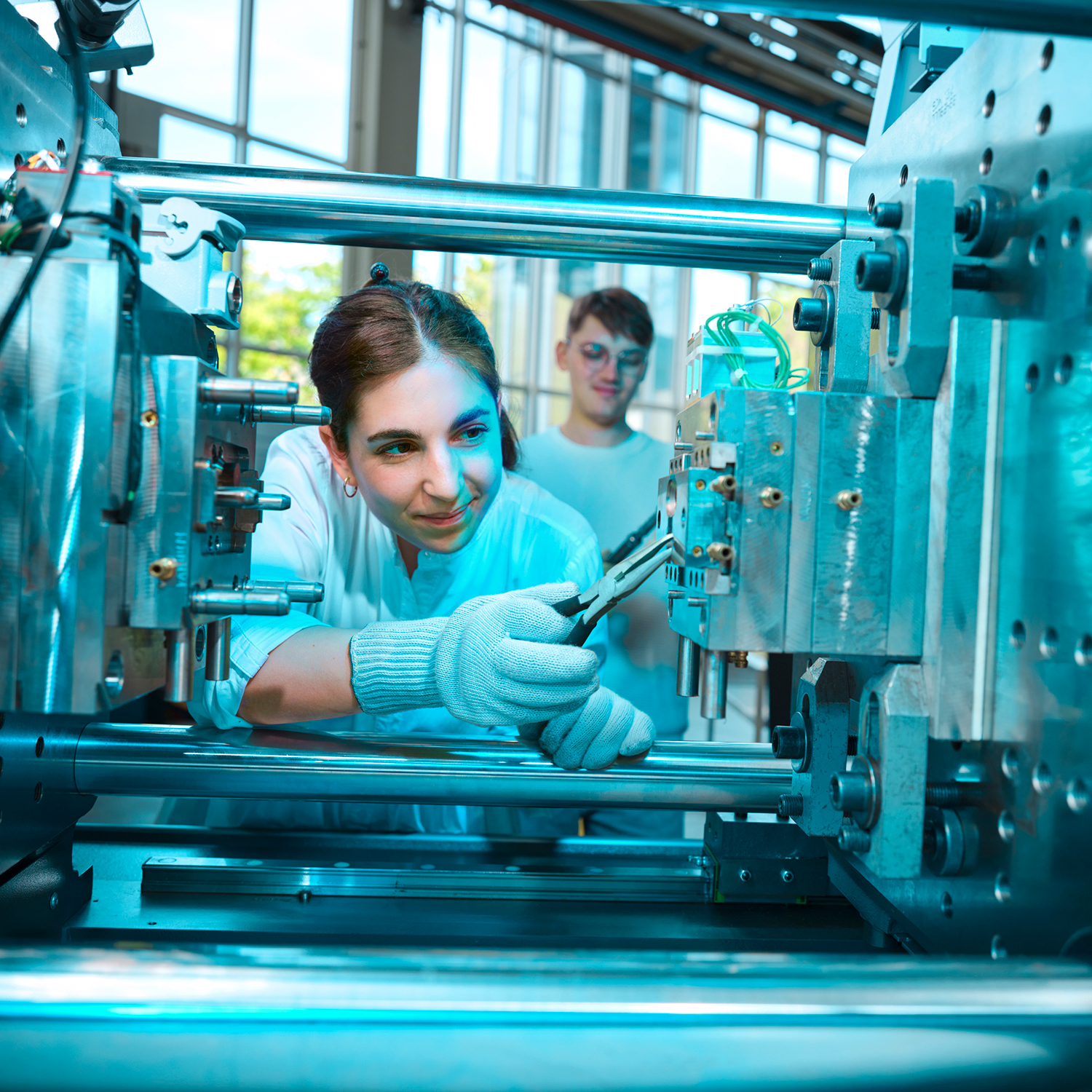 A woman stands in front of an injection molding machine and checks the machine with a tool.