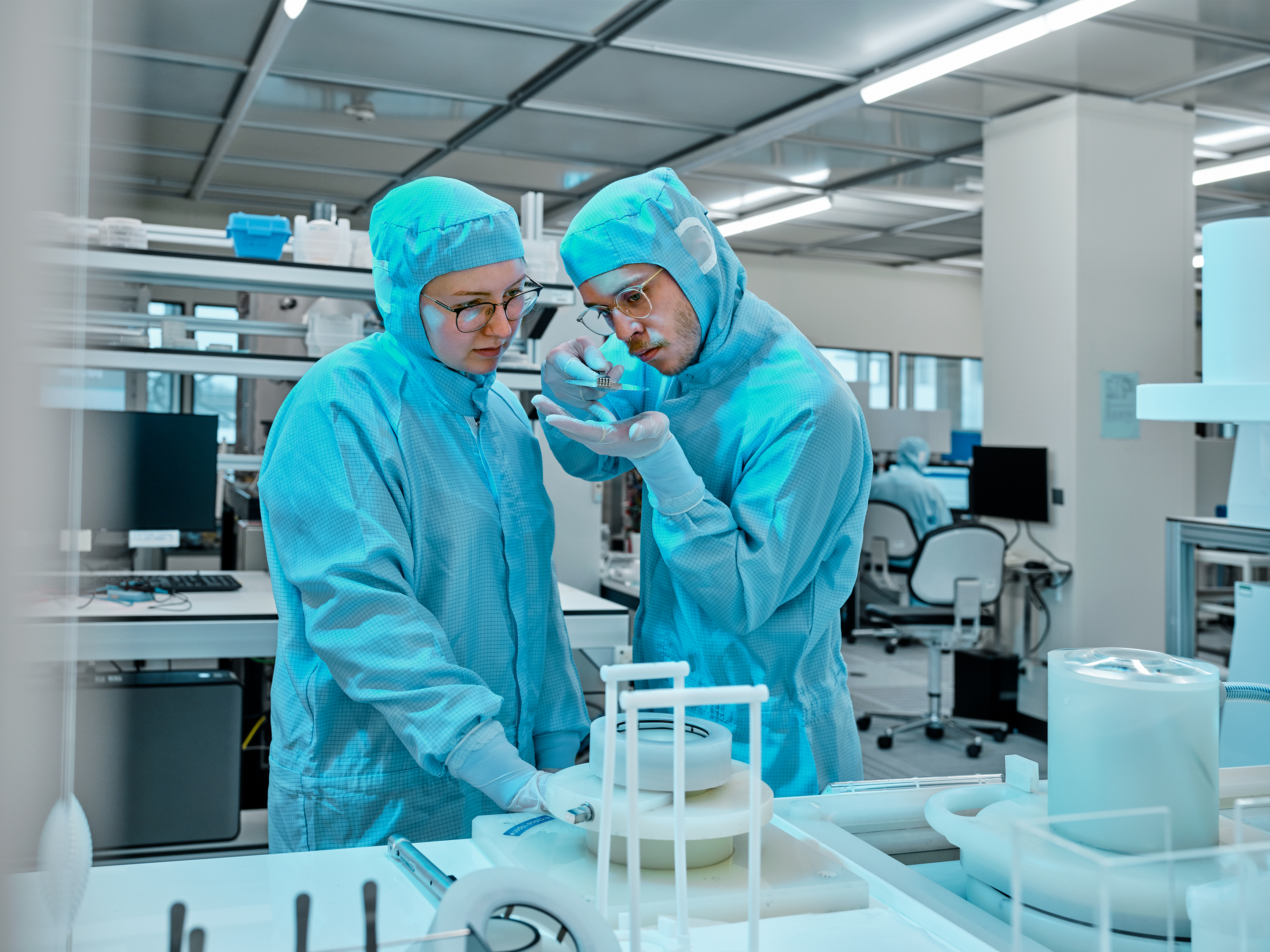 A woman and a man in protective suits stand in a laboratory, focussing at a small object in the man&#39;s hand.