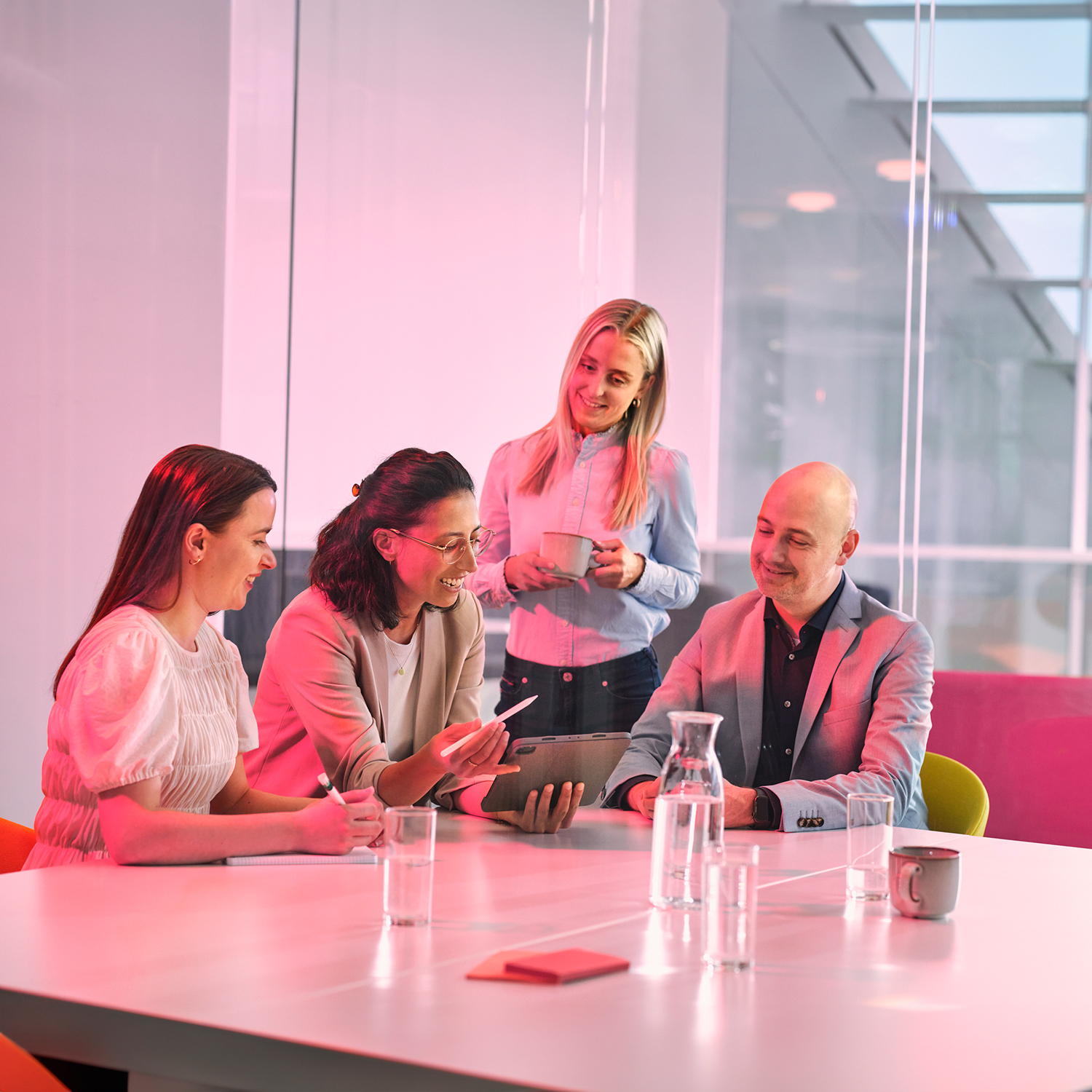 A man and two women, one with a tablet in her hand, sit at a meeting table. Another woman is standing behind them.