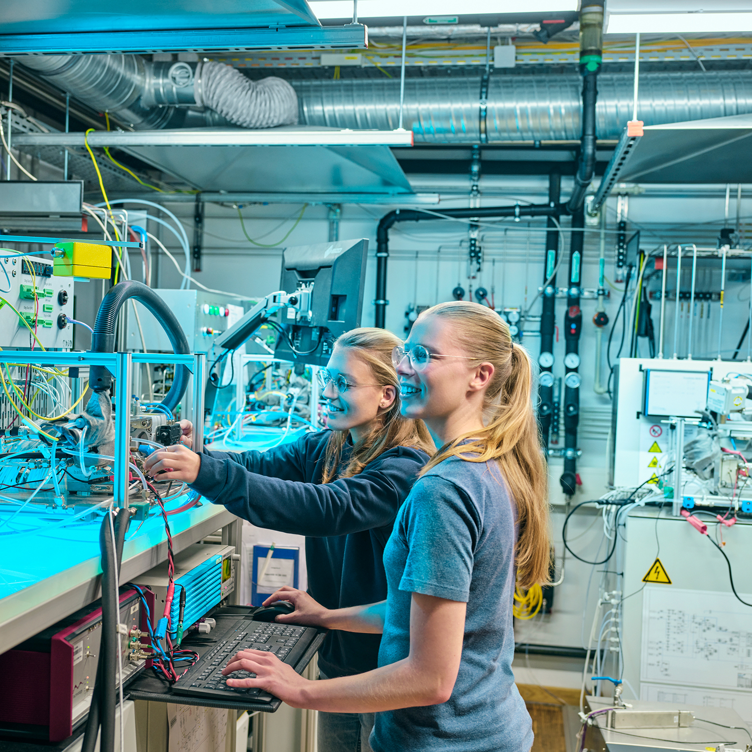 Two young women are standing in a fuel cell laboratory working on a PC. 