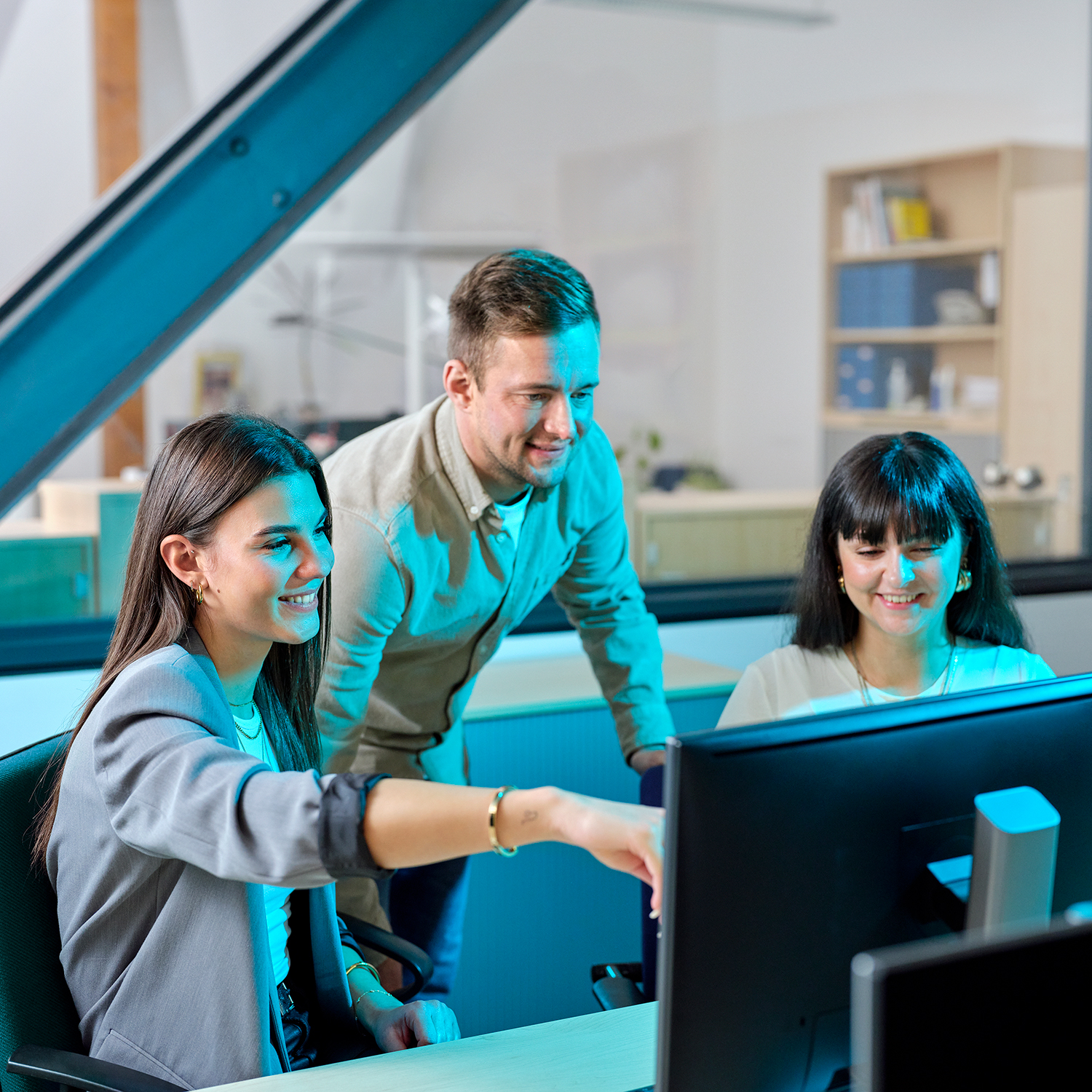 Two young women sit at a desk and look at a computer screen. A man stands in the middle and also looks at the screen.