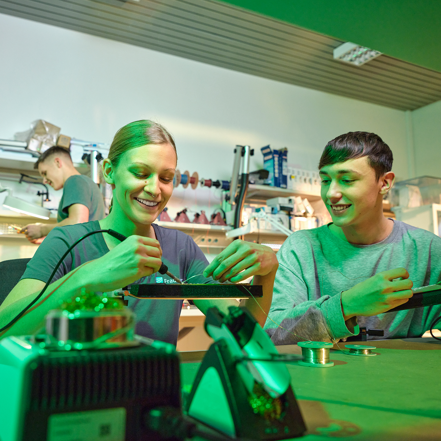 A young woman tests a circuit board with a curve tester, a young man stands behind her and watches.