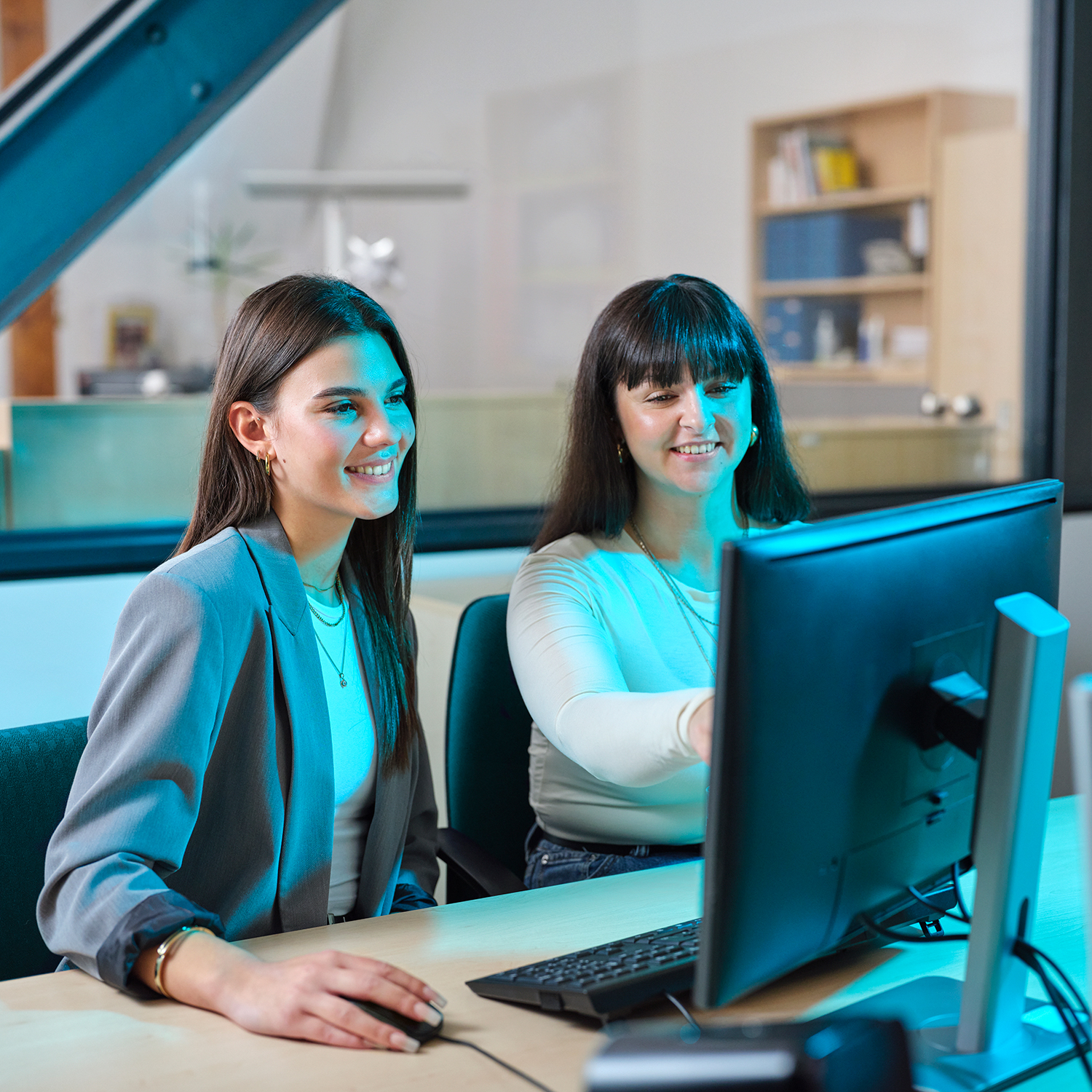 Two young women sit at a desk and look at a computer screen.