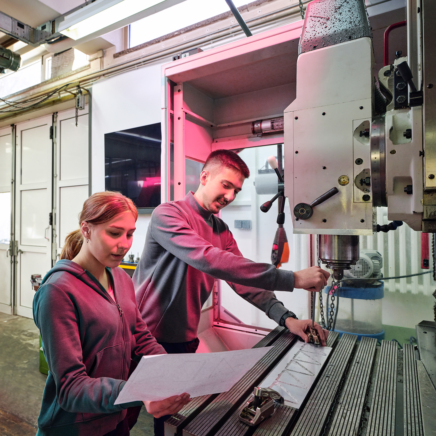 A young female and a young male trainee operate a milling machine in a workshop.