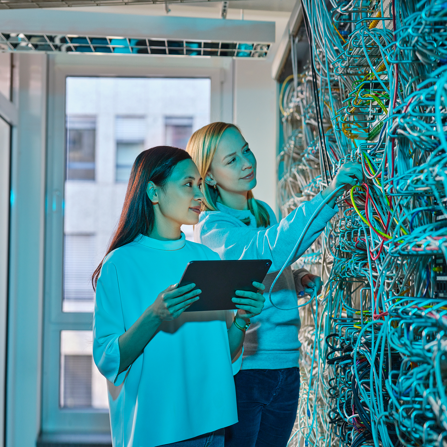 Two young women stand in front of a server rack. One woman holds a tablet, the other is checking one of the server cables.