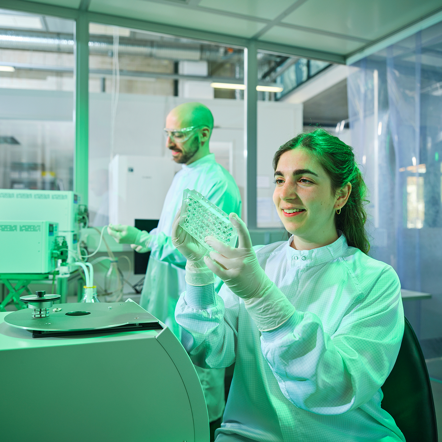 A young woman in a lab coat sits in front of laboratory device in a clean room. In the background, a man in a lab coat stands at another piece of laboratory device.