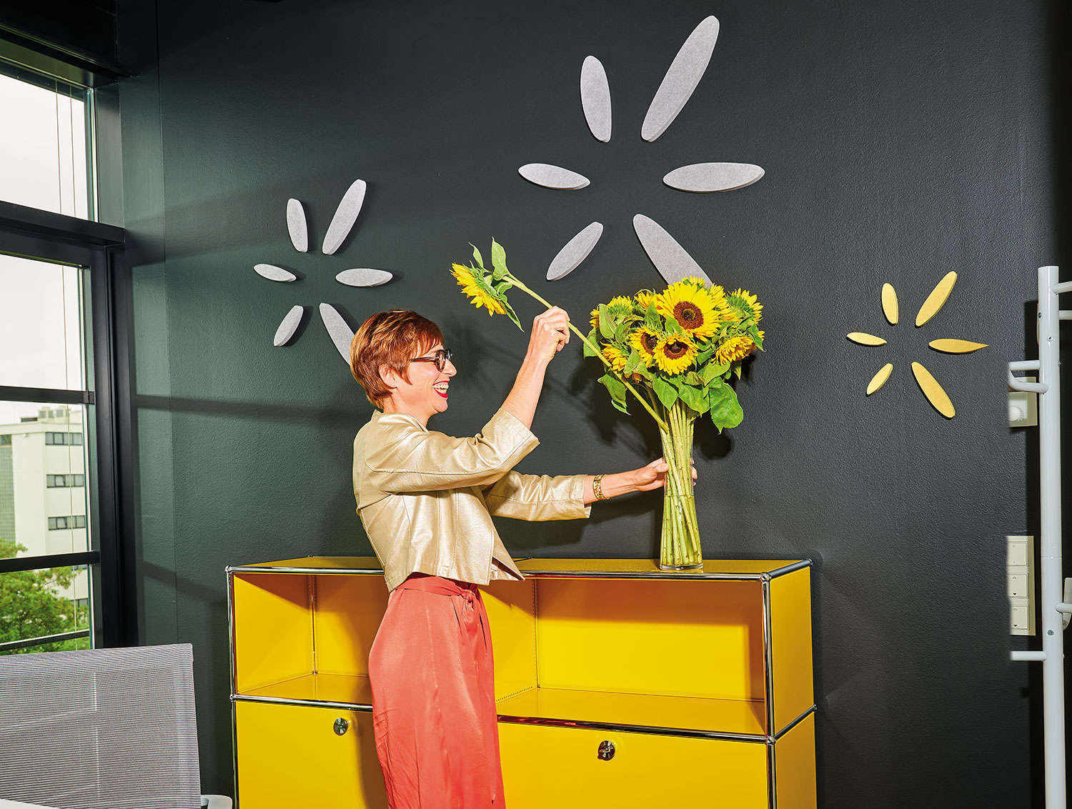 Amandine Perez in einem Raum steht vor einer gelben Kommode, sie steckt eine Sonnenblume in eine Vase mit einem Strauß Sonnenblumen.